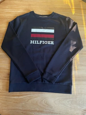 Svart sweatshirt från Hilfiger - Snygg svart sweatshirt från Hilfiger med tryckt logga och färgblock i vitt, rött och svart på bröstet. Tröjan har rund halsringning, långa ärmar och ribbade muddar vid ärmslut och nederkant. Perfekt för en avslappnad och stilren look.