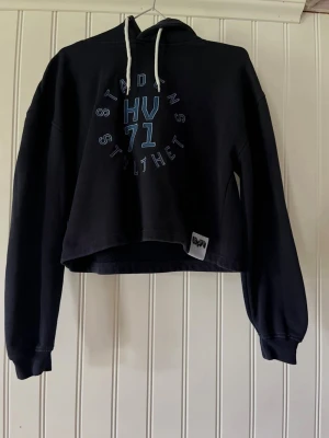 Svart cropped hoodie HV71 - Snygg svart cropped hoodie med huva och vita dragsnören. Tryck på bröstet med texten HV71 och STADENS STOLTHET i blått och vitt. Långa ärmar med muddar och en liten etikett nertill. Perfekt till ett par jeans.
