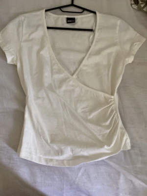 Vit omlottopp från Gina Tricot - Snygg vit omlottopp från Gina Tricot i mjuk bomull. Toppen har korta ärmar, v-ringning och lätt rynkade detaljer i sidan för en smickrande passform. Perfekt till jeans eller kjol för en fräsch look.