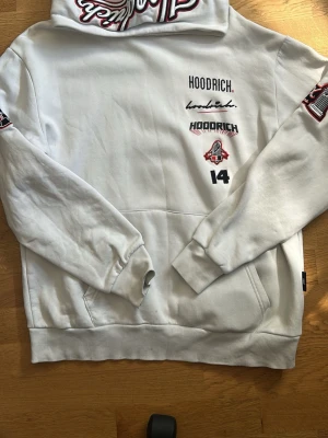 Vit hoodie från Hoodrich med tryck - Säljer en vit hoodie från Hoodrich med flera tryck i svart och rött på bröstet, ärmarna och ryggen. Hoodien har huva, känguruficka och ribbade muddar. Materialet är mjukt och skönt, perfekt för en avslappnad streetwear-look.