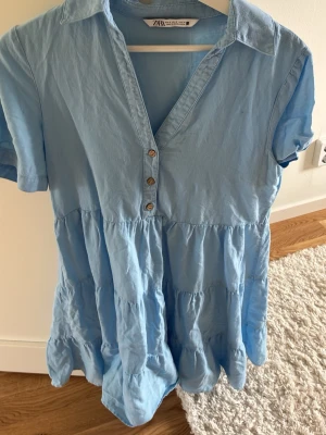 Ljusblå kort klänning från Zara - Söt ljusblå klänning från Zara med kort ärm, v-ringning och knappar framtill. Klänningen har volangdetaljer och är tillverkad i ett mjukt bomullstyg. Perfekt för varma dagar och enkel att styla med sneakers eller sandaler.