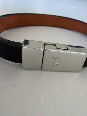 Läderarmband med magnetlås - Stilrent armband i svart läder med insida i brunt och ett rektangulärt magnetlås i blank metall. Spännet har en diskret gravyr på ovansidan. Perfekt accessoar för en modern look.