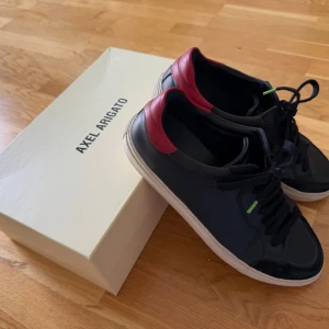 Svarta sneakers från Axel Arigato - Snygga svarta sneakers från Axel Arigato med röda detaljer vid hälen och vita sulor. Skorna har klassisk snörning och en stilren design med små gröna accenter. Perfekta för dig som vill ha en clean och modern look. Skorna är i använt men gott skick. 