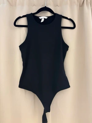 Svart body från H&M - En stilren svart body från H&M med hög halsringning och ärmlös design. Tillverkad i ett stretchigt material som sitter snyggt på kroppen. Perfekt att matcha med jeans eller kjol för en trendig look.