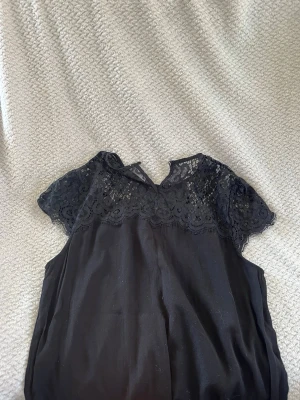 Svart blus med spetsdetaljer - Snygg svart blus med korta ärmar och vacker spets upptill. Blusen har rund halsringning och transparent spets över axlar och bröst, vilket ger en elegant look. Perfekt att styla med jeans eller kjol för en trendig outfit.