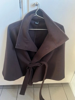 Mörkbrun kappa med knytskärp - Stilren mörkbrun kappa från Gina Tricot med bred krage och knytskärp i midjan. Kappan har en rak passform och är tillverkad i ett mjukt tyg som ger en snygg siluett. Perfekt för dig som vill ha en enkel men trendig look.