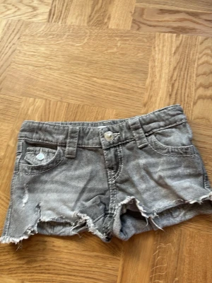Grå jeansshorts med slitna detaljer - Säljer ett par grå jeansshorts med råa, fransiga kanter och slitningar framtill. Shortsen har fickor både fram och bak, där bakfickorna har snygga broderade detaljer i blått och vitt. Klassisk knappstängning och bälteshällor.