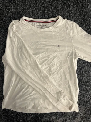 Vit beige  långärmad topp Tommy Hilfiger - Klassisk vit beige långärmad topp från Tommy Hilfiger med rund halsringning och diskret logga på bröstet. Tillverkad i mjuk bomull som känns skön mot huden. Perfekt basplagg för alla tillfällen.