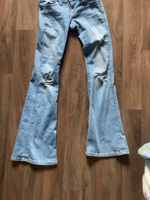Ljusblå bootcut jeans med slitningar (är designat med hål) - Säljer ett par ljusblå bootcut jeans med snygga slitningar på båda benen. Jeansen har klassisk femficksmodell och är tillverkade i denim med lite stretch. Perfekta för en avslappnad och trendig look.
