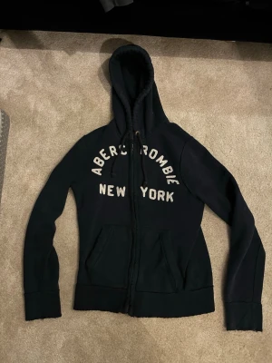 Svart hoodie Abercrombie & Fitch - Säljer en svart hoodie från Abercrombie & Fitch med dragkedja och stor huva. På bröstet står det 'Abercrombie New York' i vita bokstäver. Hoodien har kängurufickor och ribbade muddar. Perfekt för en chill och avslappnad stil.