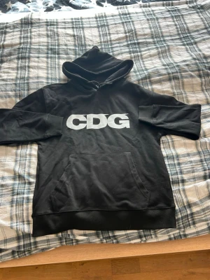 Svart CDG hoodie med tryck - Säljer en svart hoodie med stor vit CDG-logga på bröstet. Hoodien har huva med dragsko och en klassisk magficka. Tillverkad i mjukt bomullsmaterial som är skönt att bära. Perfekt för dig som gillar streetwear och enkel stil.