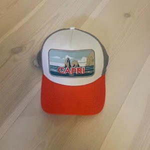 KEPS | NEW ERA. - Snygg truckerkeps med vit front, röd böjd skärm och grått mesh-tyg bak. Framtill finns ett Capri-motiv med hav och klippor samt texten 'CAPRI ITALY'. Perfekt för dig som gillar retrostil och vill sticka ut med färg. Snapback justering baktill.