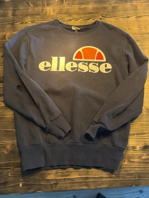 Mörkblå sweatshirt från Ellesse - Mörkblå sweatshirt från Ellesse med stort vitt och orange logotryck på bröstet. Tröjan har rund halsringning, ribbade muddar vid ärmslut och nederkant samt en avslappnad passform. Perfekt för en sportig och casual stil.