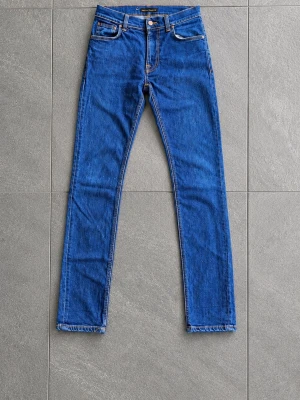 Blå raka Thin Finn jeans från Nudie Jeans - Snygga blå jeans från Nudie Jeans med klassisk rak passform och hög midja. Jeansen har fem fickor, kontrastsömmar och Nudie-logga på bakfickan. Tillverkade i slitstark denim, perfekta för en clean och tidlös look.