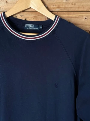 Vintage Polo Ralph Lauren 90s kortärmad sweatshirt navy XL - Vintage 90-tals Polo by Ralph Lauren kortärmad sweatshirt i mörkblå bomull. Ovanlig modell med raglanärm, sweatshirtkvalitet, vinröda/vita ribbade detaljer och broderad Polo Pony.  Fint vintagebruksskick med lätt naturlig patina.  Storlek XL | 100 % bomull | Made in Hong Kong  Bröstvidd 59 cm | Ärmlängd från hals 40 cm | Längd 73 cm