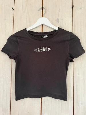 Svart croppad t-shirt från Divided - Snygg svart croppad t-shirt från Divided med vit text 'OREGON' på bröstet. T-shirten har rund halsringning och korta ärmar. Perfekt till jeans eller kjol.