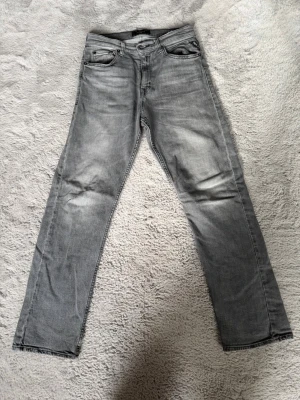 Grå raka jeans med slitningar - Säljer ett par gråa replay jeans med raka ben och diskreta slitningar vid knäna. Jeansen har klassisk femficksdesign och detaljerade sömmar på bakfickorna. Materialet är denim i bomull med en mjuk tvättad finish. Perfekta för en avslappnad och trendig look.
