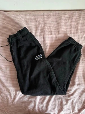 Svarta fleece mjukisbyxor med patch - På Svarta fleecebyxor från Junkyard.  Mjuka och bekväma, perfekta för en avslappnad look.  Har ett gummiband i midjan för att justera passformen, men plastknappen har gått sönder lite (syns i bilden) Detta påverkar inte byxorna i övrigt och de går fortfarande att använda som vanligt.  I fint skick för övrigt.