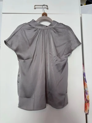 Grå satinblus med korta ärmar - Stilren grå blus i glansigt satinmaterial med korta ärmar och lätt rynkad detalj vid halsen. Blusen har en avslappnad passform och en mjuk, skön känsla. Perfekt att matcha med jeans eller kjol för en trendig look.