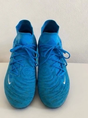 Blå Nike fotbollsskor med högt skaft - Säljer ett par blå Nike fotbollsskor med högt skaft och snörning. Skorna har en vit Nike-logga på sidan och framtill samt mönstrad ovansida för bättre bollkontroll. Perfekta för dig som vill ha stilrena och funktionella skor på planen.