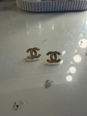 Guldiga örhängen med stenar - Snygga örhängen i guldfärgad metall med gnistrande stenar formade som två sammanlänkade C:n. Perfekta för dig som gillar klassisk och elegant stil med en touch av lyx.