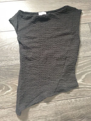 Svart asymmetrisk topp Van Der Rock - Trendig svart topp från Van Der Rock med asymmetrisk nederkant och strukturerad yta. Toppen är ärmlös och har en tight passform som framhäver siluetten. Perfekt för dig som gillar en edgy och modern stil.