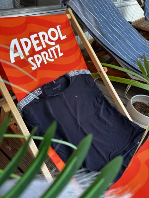 Mörkblå t-shirt från Tommy Hilfiger - Snygg marinblå t-shirt från Tommy Hilfiger med logga på bröstet och märkestext längs kragen. Klassisk passform och mjukt bomullsmaterial som passar perfekt till sommaren. Enkel att matcha med shorts eller jeans.