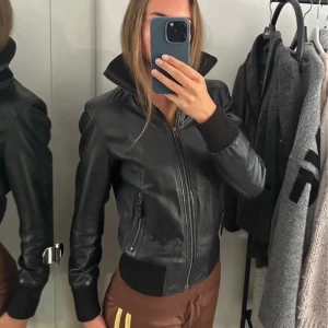 Svart skinnjacka med ribbade detaljer - Snygg svart skinnjacka med dragkedja framtill, ribbade muddar vid ärmslut och midja samt hög krage. Jackan har en klassisk bomberstil och två fickor med dragkedja på sidorna. Perfekt för dig som vill ha en tidlös och cool look.