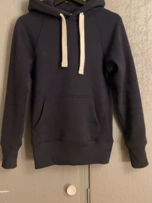 Russemerch hoodie - Mysig mörkblå hoodie med huva och vita dragsnören. Klassisk känguruficka framtill och ribbade muddar vid ärmslut och nederkant. Perfekt för en avslappnad stil och enkel att matcha med jeans eller joggers.