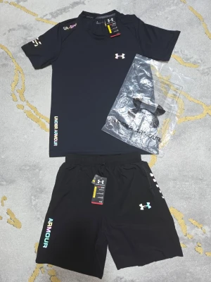 Svart träningsset från Under Armour - Snyggt svart träningsset från Under Armour med t-shirt och shorts. Båda plaggen har färgglada loggor och textdetaljer. Tillverkade i ett lätt och snabbtorkande material som passar perfekt för gym eller löpning.