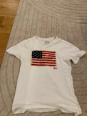 Vit t-shirt med amerikansk flagga Polo - Vit t-shirt från Polo Ralph Lauren med tryck av amerikanska flaggan på bröstet och diskret RL-brodyr. Klassisk rund hals och korta ärmar. Perfekt för en avslappnad och cool stil.