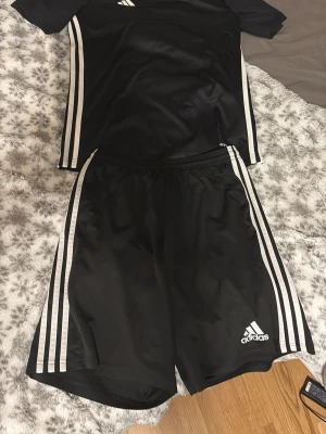 Svart Adidas träningsset med vita ränder - Säljer ett svart träningsset från Adidas med klassiska vita ränder längs sidorna. Setet består av en t-shirt och shorts i lätt och snabbtorkande material, perfekt för träning eller match. Adidas-logga på både tröja och shorts. Snygg och sportig look!