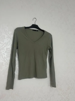 Olivgrön ribbad långärmad topp - Snygg olivgrön långärmad topp med v-ringning och ribbad struktur. Perfekt att styla med jeans eller kjol för en enkel men trendig look. Materialet känns mjukt och stretchigt, vilket gör den bekväm att bära hela dagen.