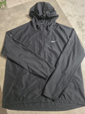Svart Nike vindjacka med huva - Snygg svart vindjacka från Nike med huva och dragkedja framtill. Jackan har två sidofickor med dragkedja och reflekterande Nike-logga på bröstet. Tillverkad i lätt och tunt material, perfekt för löpning eller promenader. Sportig och stilren design.