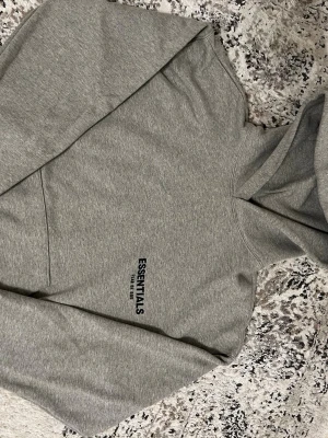 Grå Essentials Fear of God set - Säljer en grå hoodie från Essentials Fear of God med svart logga på bröstet och stor text på ryggen. Klassisk passform med huva och känguruficka framtill. Tillverkad i mjukt sweatshirtmaterial som är perfekt för chill dagar.