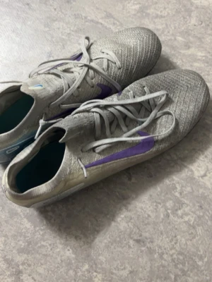 Grå sportskor med lila swoosh - Snygga sportskor i grått med lila swoosh på sidan och blå detaljer på sulan. Ovandelen är i mesh-liknande material för bra andningsförmåga och skorna har klassisk snörning. Perfekta för dig som gillar sportig stil och vill sticka ut med färgade detaljer.