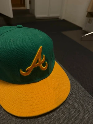 Grön och gul New era Keps - Snygg keps i grön färg med gul skärm och en stor gul broderad A-logga framtill. Klassisk snapback-modell med platt skärm och ventilationshål på ovansidan. Perfekt för dig som vill sticka ut med färg och stil.