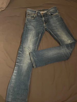 Grim trim nudie jeans - Snygga blå jeans från Nudie Jeans med klassisk femficksdesign och orangea sömmar. Jeansen har en rak passform och är tillverkade i slitstarkt denim med coola slitningar och detaljer på bakfickorna. Perfekt för dig som gillar en tidlös och avslappnad stil.