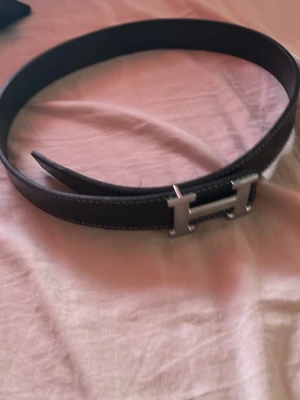 Svart bälte med silverfärgat H-spänne - Snyggt svart bälte med ett stort H-format spänne i silverfärg. Bältet har en stilren design med vita sömmar längs kanterna och passar perfekt till jeans eller byxor för en trendig look.
