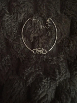 Silverfärgat armband med hjärtdetalj - Säljer ett stilrent armband i silverfärg med en snygg hjärtdetalj i mitten. Armbandet har en tunn kedja med justerbar längd och enkel karbinhake. Perfekt accessoar för att lyfta din outfit.