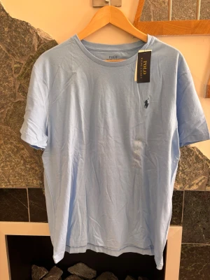 Ljusblå t-shirt från Polo Ralph Lauren, strlk L - Snygg ljusblå t-shirt från Polo Ralph Lauren med klassisk passform och det ikoniska logobroderiet på bröstet. Tillverkad i mjuk bomull och har rund halsringning samt korta ärmar. Perfekt för en avslappnad och stilren look.