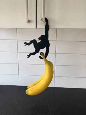 Svart bananhängare apa - Rolig bananhängare i form av en svart apa som hängs på ett skåp eller handtag. Perfekt för att förvara bananer på ett kreativt sätt i köket. Tillverkad i metall med matt finish och stilren design.