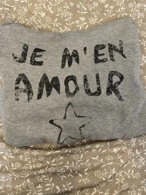 Grå hoodie - Säljer en grå hoodie med svart texten 'JE M'EN AMOUR' och en stjärna framtill. Hoodien har huva med dragsko och ribbade muddar vid ärmar och nederkant. Perfekt för en chill och avslappnad stil.