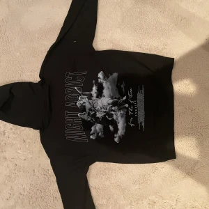 Svart hoodie från Night Addict - Snygg svart hoodie från Night Addict med tryck på baksidan. Den har en klassisk design med huva och långa ärmar. Perfekt för en avslappnad stil.