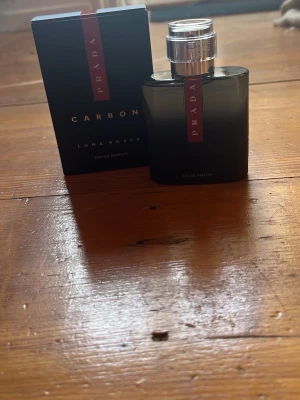 Prada Luna Rossa Carbon EdP - Prada Luna Rossa Carbon Eau de Parfum med elegant svart och grå glasflaska, silverfärgad spraykork och röd detalj med Prada-logga. Doften är modern och maskulin, perfekt för dig som gillar exklusiva parfymer. Knappt andvänd då jag inte tyckte den passa mig jag har bara sprutat en gång
