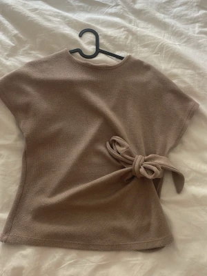 Beige stickad topp med knyt från Zara - Säljer en beige stickad topp från Zara med korta ärmar och snygg knytning i sidan. Perfekt till jeans eller kjol för en chill men ändå stilsäker look. Materialet är mjukt och toppen har en avslappnad passform.