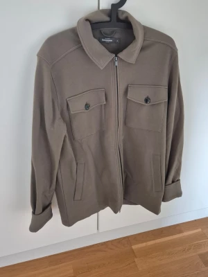 Dressman Overshirt - Stilren beige overshirt med dragkedja framtill och två bröstfickor med knapp. Overshirten har klassisk krage och raka ärmar med knapp vid ärmslut. Tillverkad i ett mjukt tyg som ger en avslappnad look.