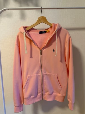 Ljusrosa kofta från Polo Ralph Lauren - Ljusrosa hoodie med dragkedja från Polo Ralph Lauren. Klassisk modell med huva och vita dragsnören, två stora fickor framtill och broderad logga på bröstet. Tillverkad i mjukt bomullsmaterial som känns skönt mot huden. Perfekt för en avslappnad och stilren look.