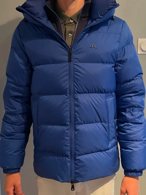Blå j.Linderberg dunjacka  - Snygg blå dunjacka med hög krage och tvåvägsdragkedja. Jackan har en diskret logga på bröstet, två sidofickor och breda, quiltade paneler. Perfekt för kalla vinterdagar och ger en modern, sportig look. Nypris 4800. Använd en vinter. Hör av dig vid frågor eller funderingar!! 