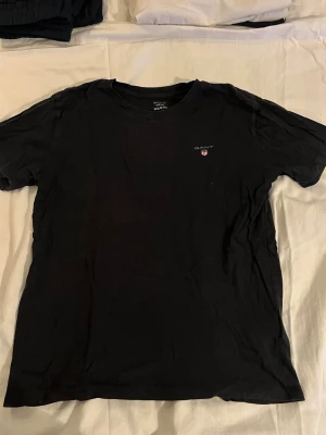 Svart t-shirt från GANT - Klassisk svart t-shirt från GANT med rund halsringning och liten logga på bröstet. Tillverkad i mjuk bomull för en bekväm känsla. Perfekt basplagg som funkar till det mesta och har en enkel, stilren design.